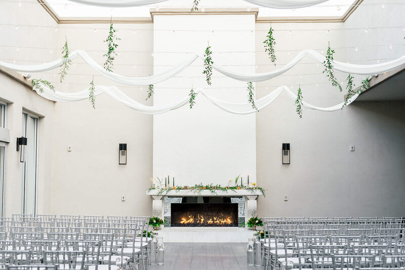 A bright and modern ceremony setup at SoHo63, a Chandler wedding and event venue, features rows of silver chairs facing a white fireplace framed with greenery and florals. Soft white drapery and hanging vines create a sophisticated and airy backdrop for a stylish Arizona wedding.