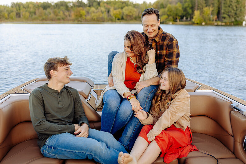 A family of four sits smiling on a boat by the water. The father and mother are in the middle, with the son on the left and the daughter on the right. Captured by a talented Minnesota photographer, trees and a clear sky create a stunning background.