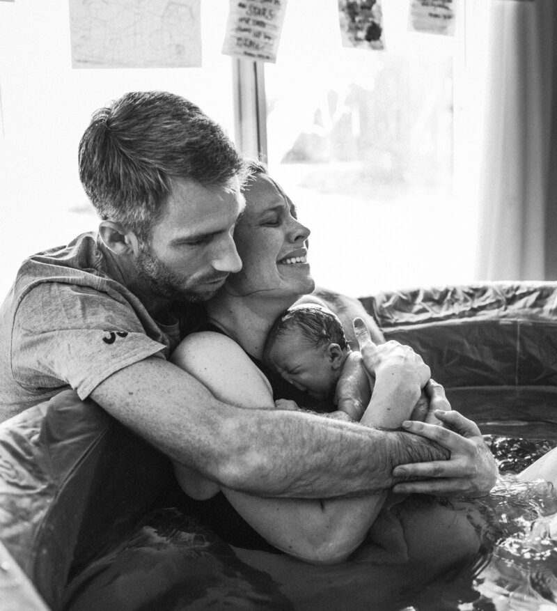 A mom is filled with emotion as she holds her just born baby who was just born and brought to her chest while int he birth tub as her husband wraps his arms around her and the newborn baby.