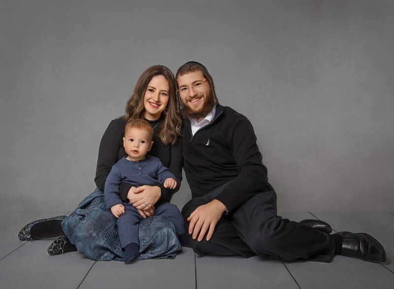 Brooklyn first birthday family photoshoot. Mom, dad, and baby boy are sitting on the ground and smiling for a first birthday photoshoot. They are wearing hues of blue. Captured by best Brooklyn, NY newborn photographer Chaya Bornstein.
