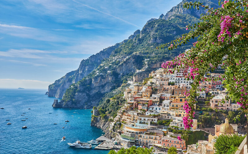 A colorful cliffside town overlooking bright blue water, with boats in the harbor and vibrant flowers in the foreground. Terraced buildings climb the steep mountainside under a clear sky.