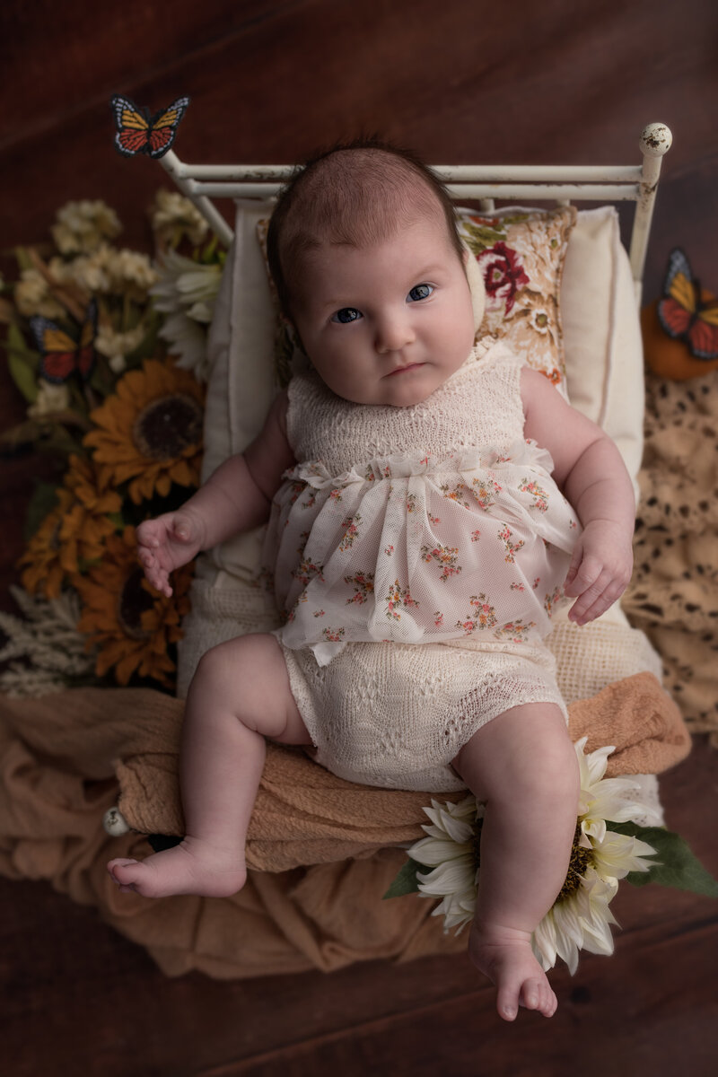 Baby girl milestone image with butterflies, floral textures, and a rustic vintage bed setting—captured in warm, earthy tones.