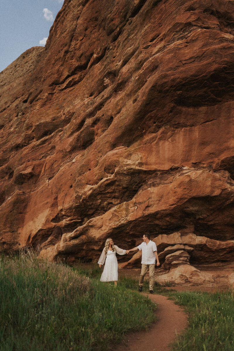 A couple holds hands and walks together along a path with a large, red rock formation towering beside them, set in a grassy field in Denver Colorado.