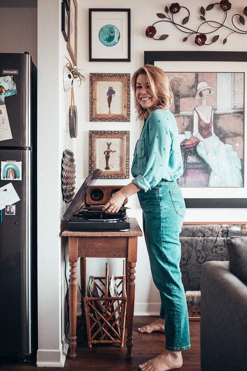 Lauren Hughes, menstrual cycle coach and cyclical living expert, smiles in a denim on denim outfit while placing a record on an open record player.