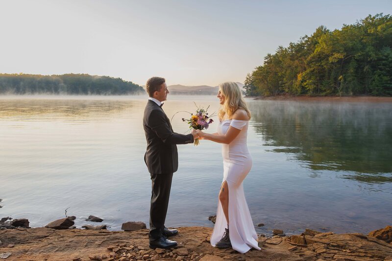 A bride and groom stand facing each other while holding hands during their elopement ceremony. The bride is wearing a fitted white gown with a thigh high slit showing off her hiking boots while the groom wears a black tux. They are holding a bouquet of wildflowers in between them. They are standing on red rocks in front of a lake while the sun rises. Steam is coming off the lake around them with a large mountain range visible in the distance behind them.