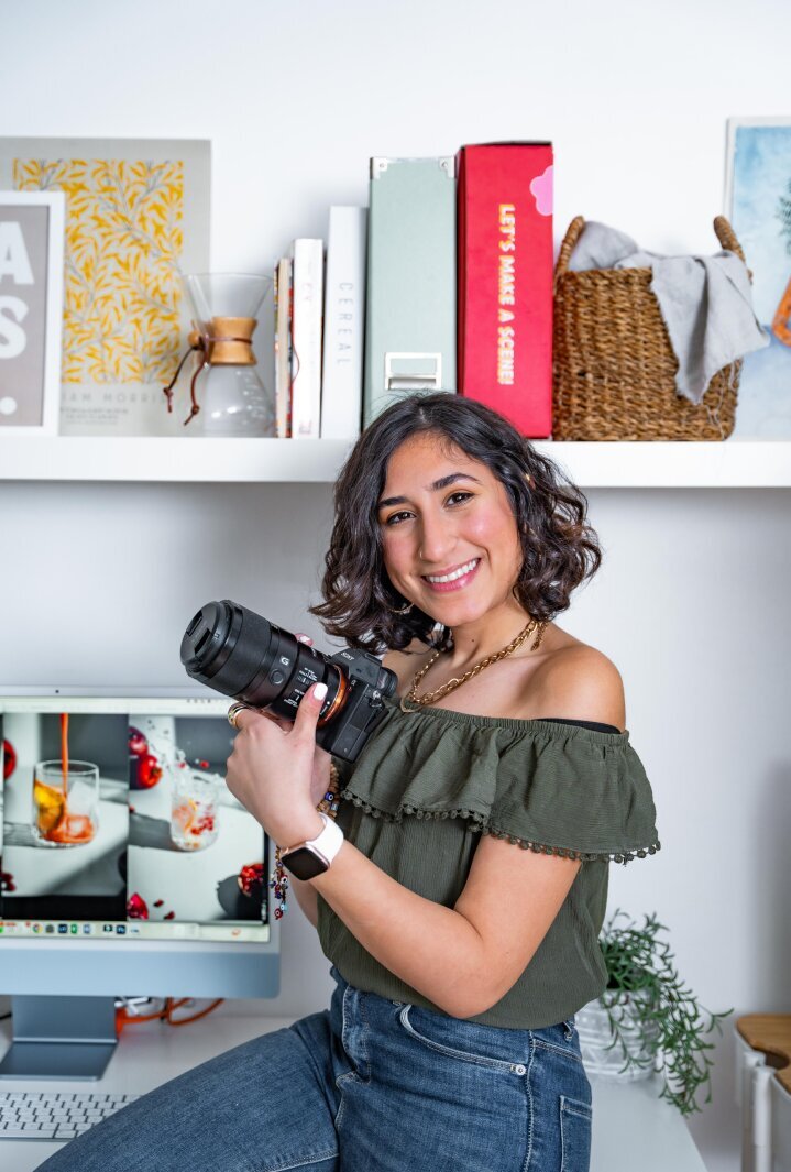 an image of Shahahd Foodie's Flare founder and lead photographer, sitting on her desk, holding her professional camera with her food photography displaying in the back on her computer screen