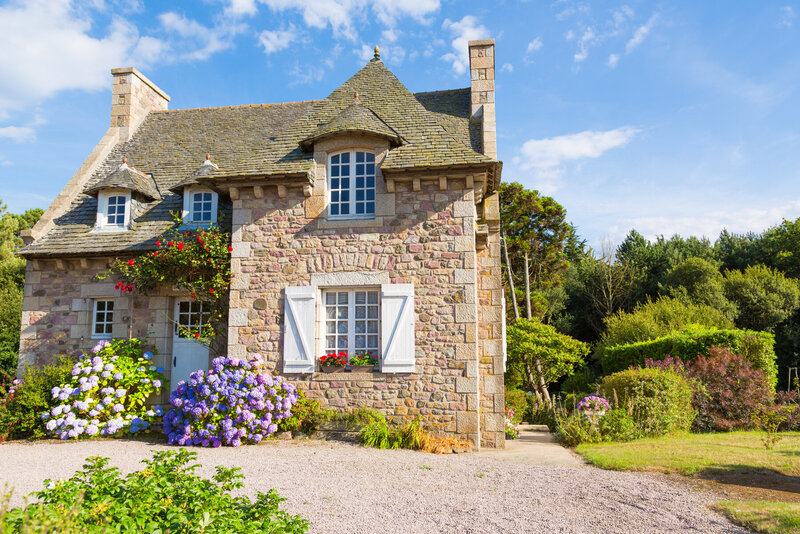 A charming stone cottage with a steep, sloped roof is surrounded by lush greenery and vibrant flowers, including purple hydrangeas. The house features white-framed windows, blue sky in the background, and a gravel path leading to the entrance.