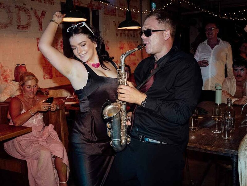 Saxophonist in sunglasses performing at a party while a guest dances beside him in a dimly lit bar venue.