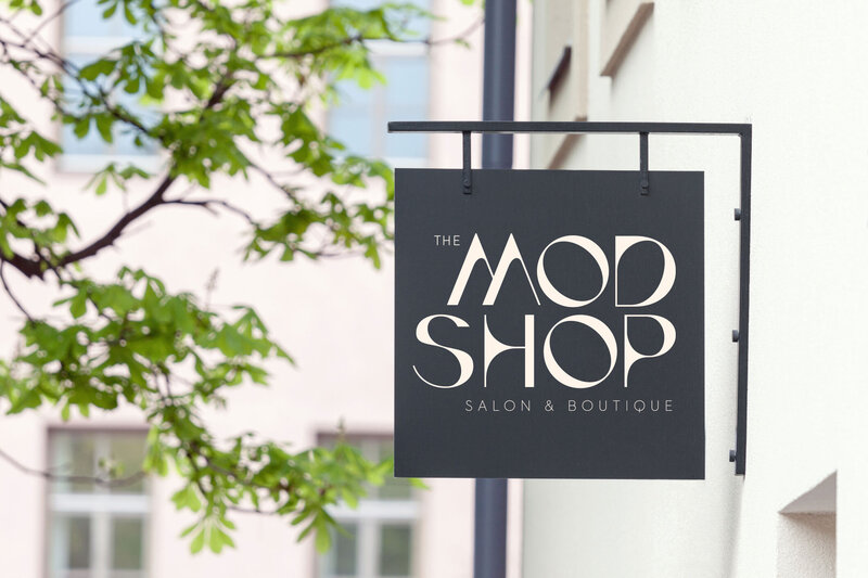 A black sign hanging up on a white outdoor wall. In white letters the sign reads "The Mod Shop Salon & Boutique".