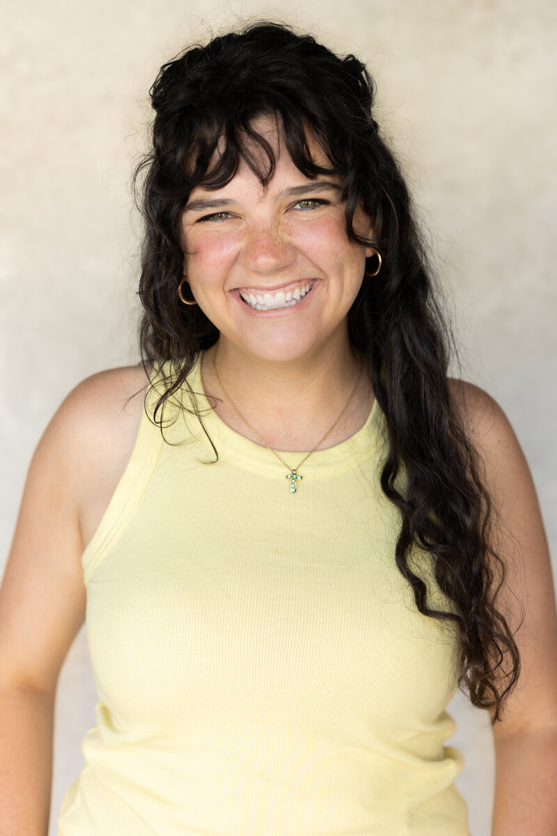 Brand photographer in Charleston, SC posing with her camera. She has dark hair and bangs and is wearing a light yellow tank top.