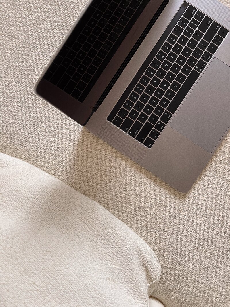 Open laptop on a textured cream-colored floor, with a black keyboard and touchpad, next to a soft cushion, conveying a cozy and minimalist vibe.