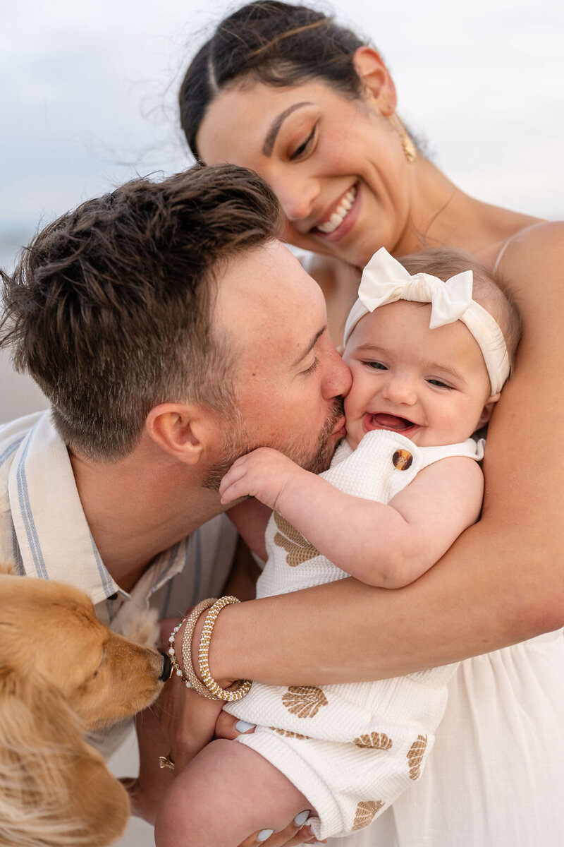 mother holds smiling baby as father kisses her cheek while holding their family dog on topsail island