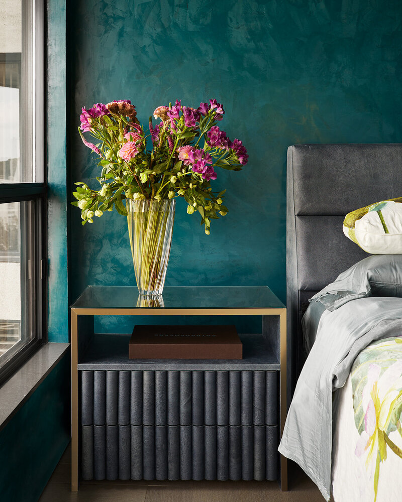 A bedside table with grey channeling and a vase of pink flowers  on top in a bedroom with grey, aqua, and green tones