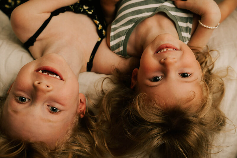 Two sisters look at the camera as they lie upside down from a bedside.