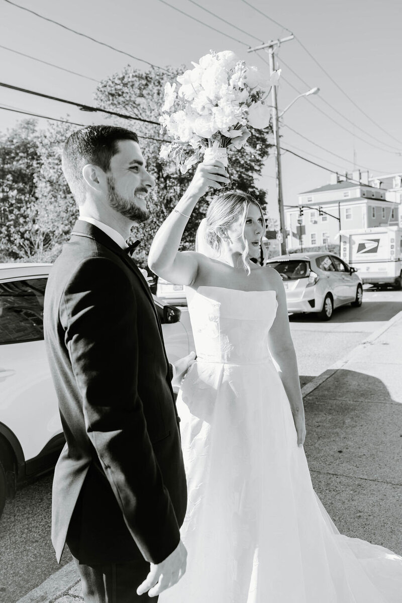 Ochre Court Newport | A bride joyfully raises a bouquet beside a groom on a sunny street, both smiling. They're surrounded by parked cars and trees.