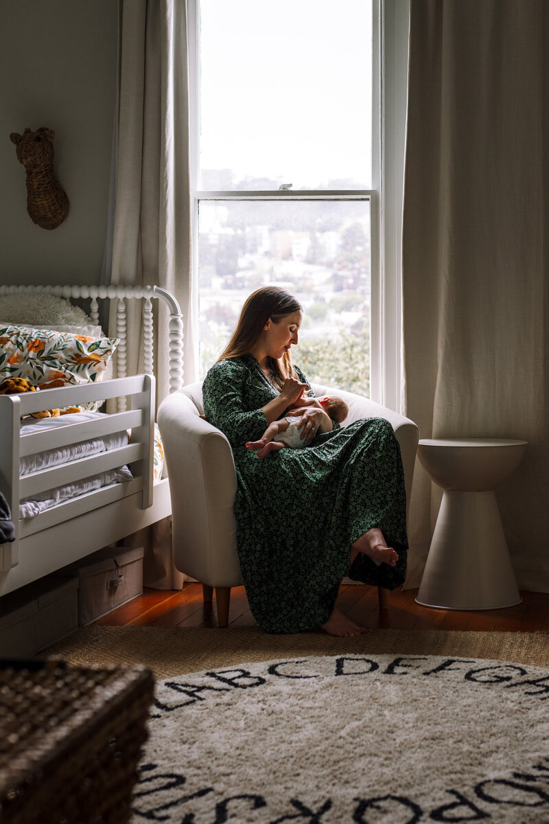 mom in green dress sitting in nursery in front of window nursing newborn