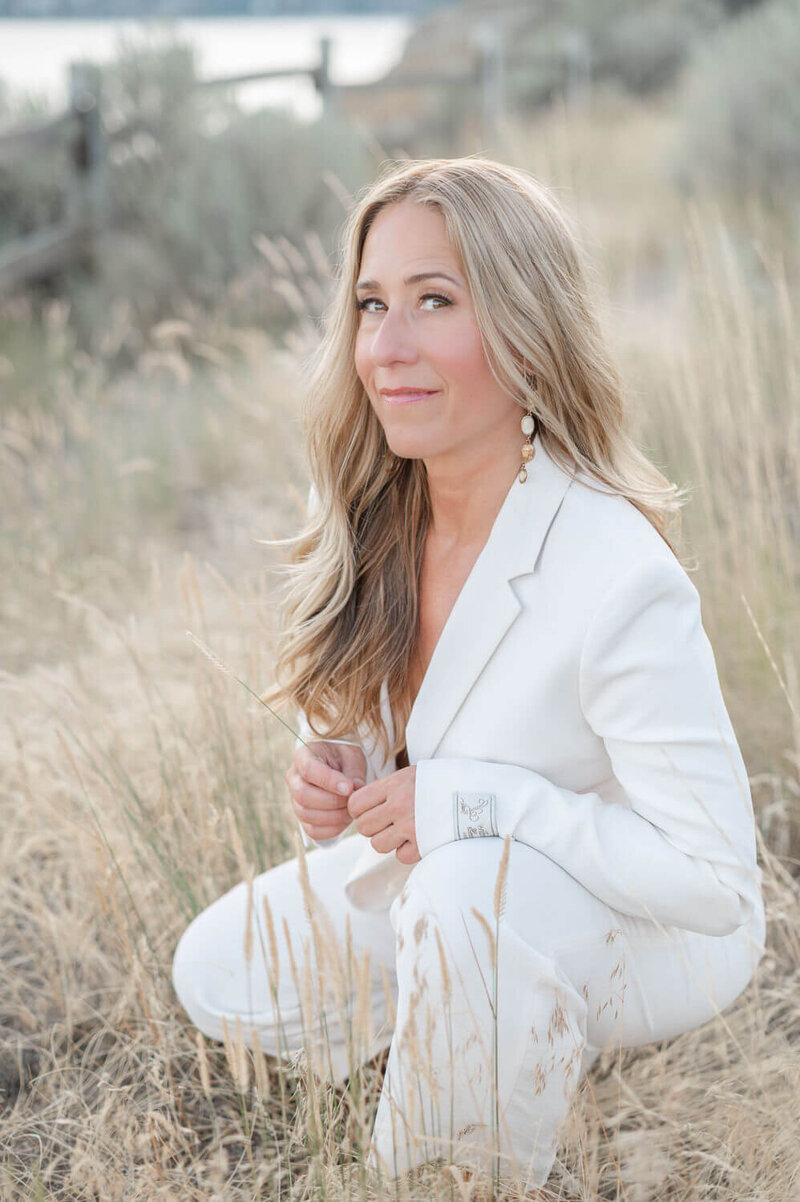 lifestyl portrait of business woman in a white suit kneeling in long grass outdoors and smiling at the camera