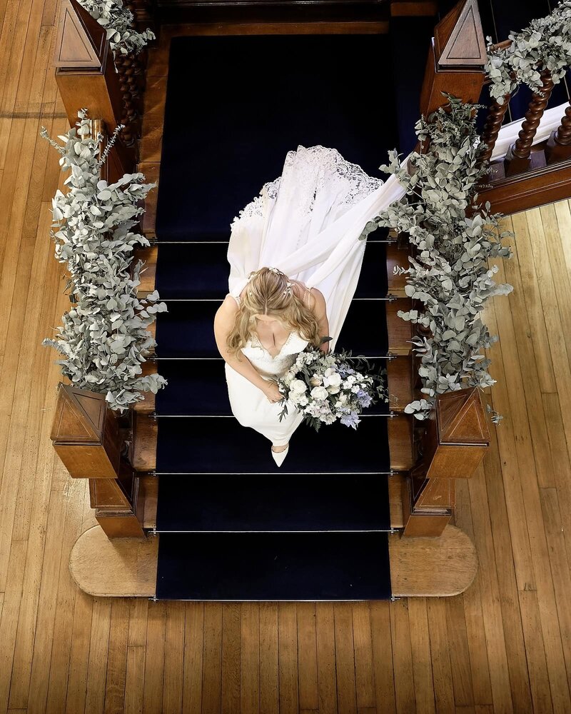 view from above as bride walks down stairs surrounded by flowers, holding her wedding bouquet