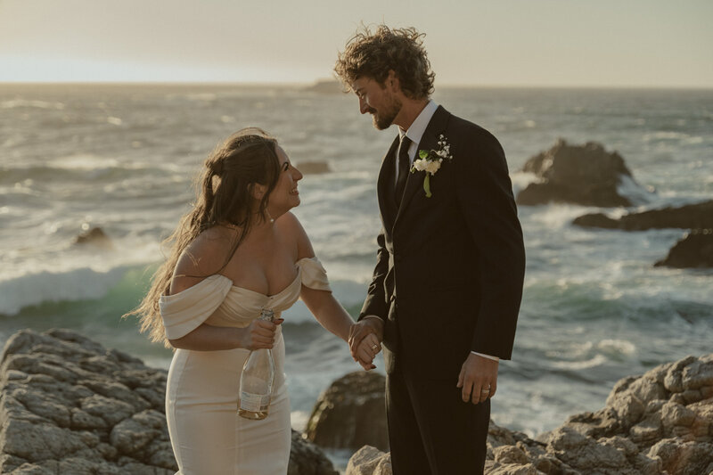 Big Sur bride and groom smiling at each other while bride holds a bottle of champagne with Big Sur waves and coastline in the background