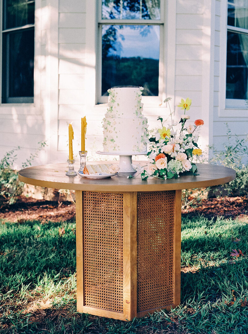 Rattan cake table with white, yellow and orange flower accent arrangement and taper candles