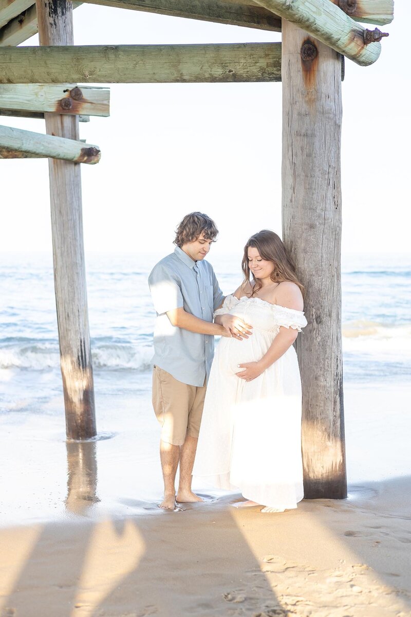Maternity Session with couple standing on a beach facing each other cradling her belly by Virginia Beach Family Photographer