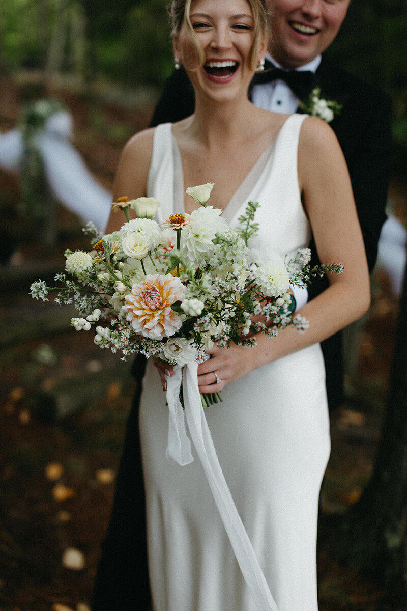 A bride in a white dress holds a bouquet of white and orange flowers, laughing with her groom in a black suit partially visible behind her at a Muskoka cottage wedding by Toronto wedding photographer, Jennifer van Son.