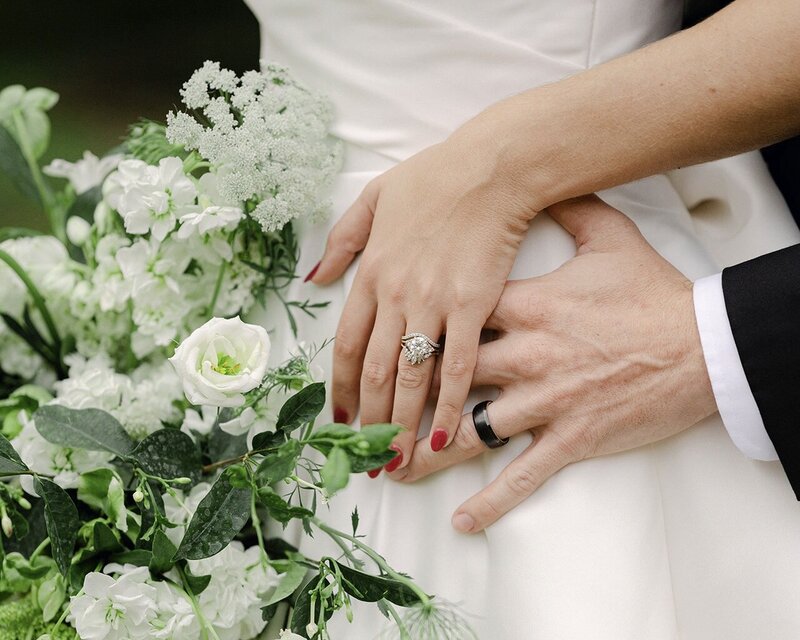 Close up photograph of bride and groom holding hands near a bouquet to show off their new wedding rings.