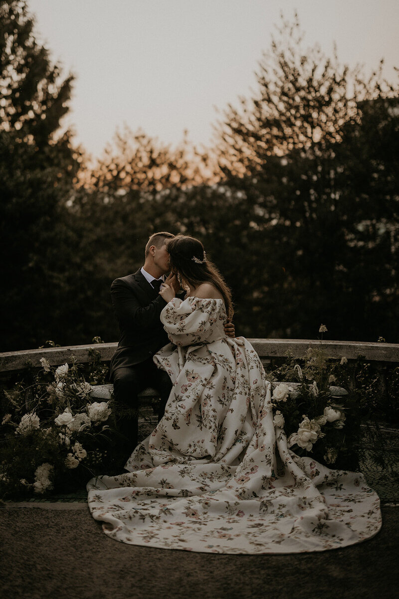 A couple shares a kiss outdoors at sunset, with the bride in a floral gown.