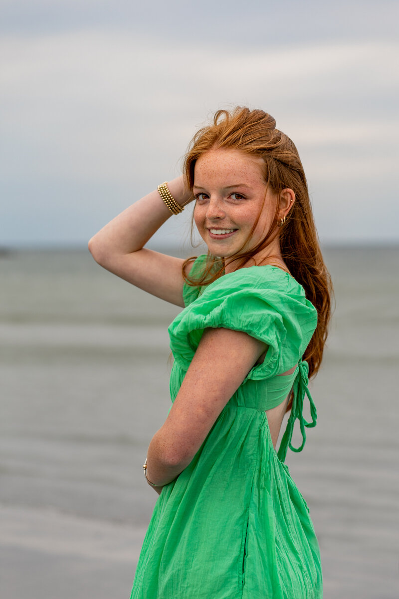 Photo of senior girl on the beach looking over her shoulder while holding hair back, and smiling at the camera.