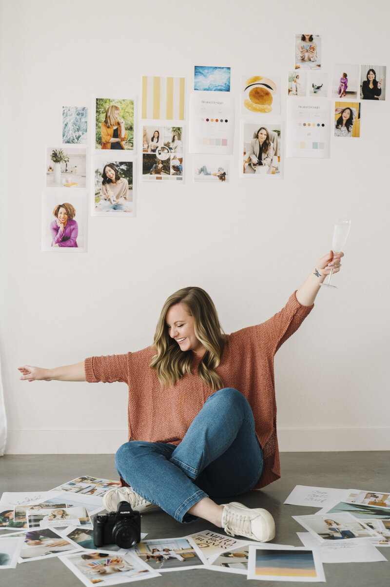 Krista Marie, sitting on the ground with her camera and printed brand photos, holding up a glass of champagne while looking away from the camera