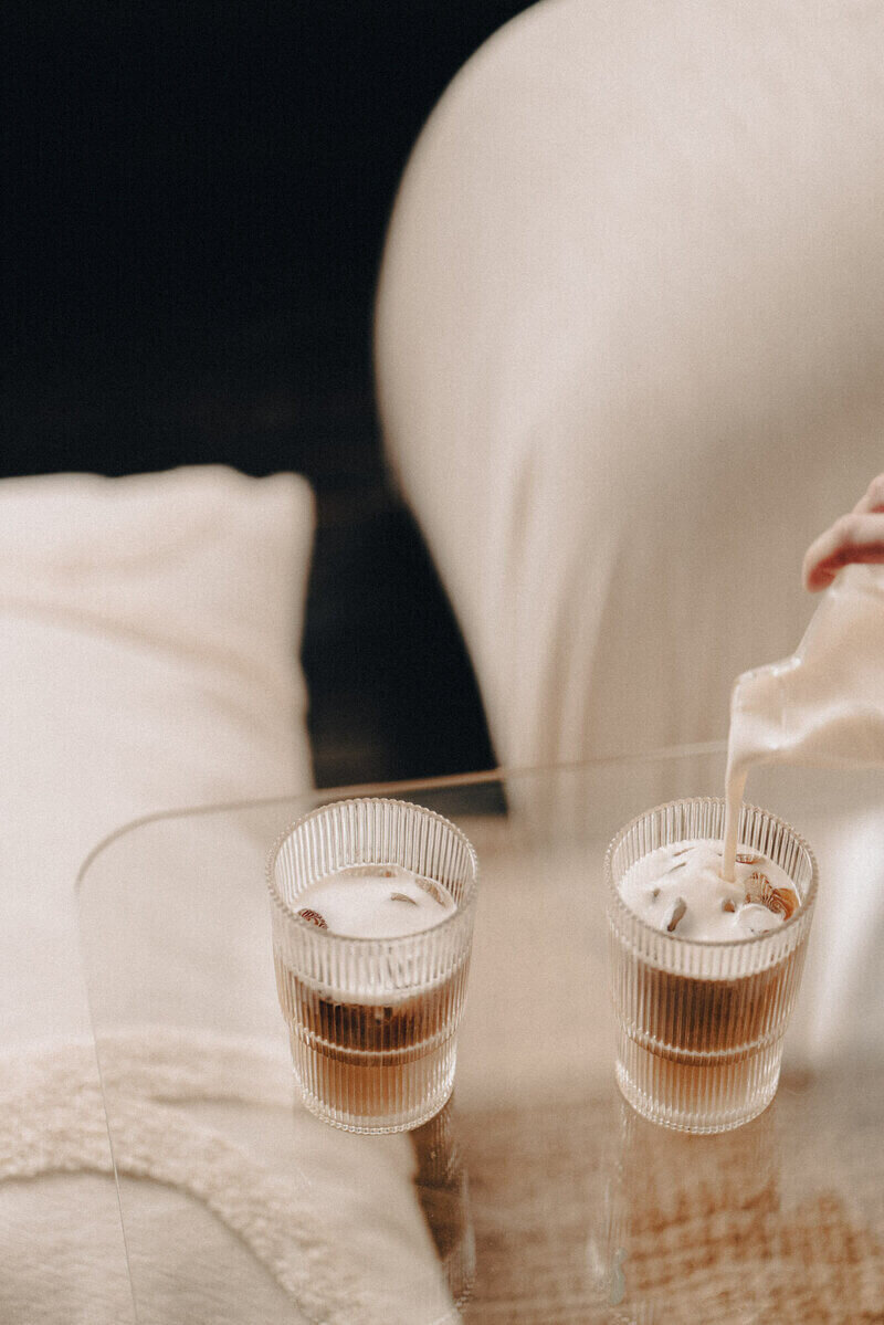 A hand pours milk into a glass of coffee, blending the two liquids together beautifully.