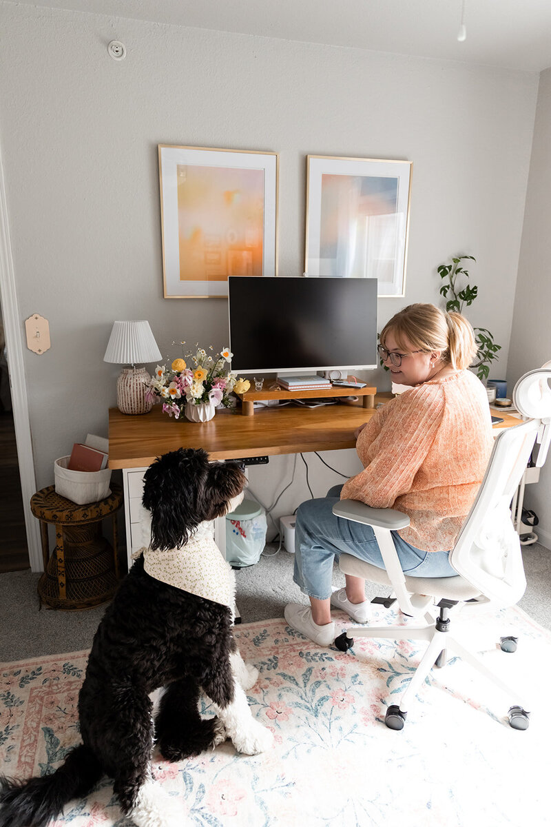 Woman with blonde hair in claw clip wearing pink blouse sitting at desk looking at black and white dog