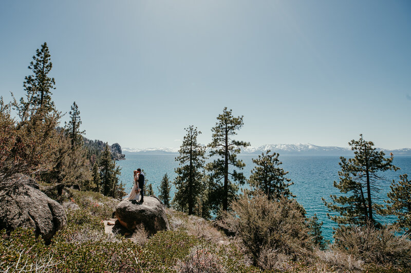 Bride and groom standing on a large rock at Logan Shoals in Lake Tahoe, surrounded by pine trees and overlooking the deep blue lake with snow-capped mountains in the distance.