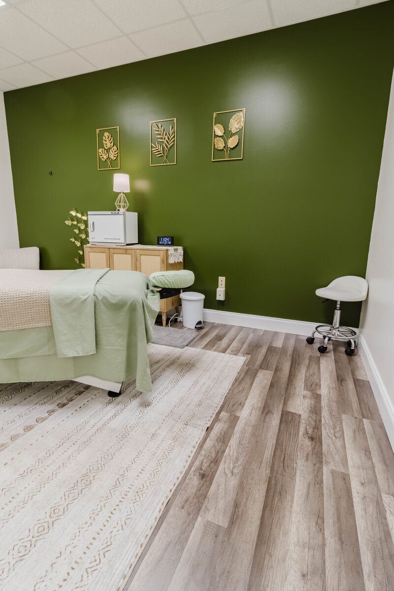 Modern massage therapy room with a calming green accent wall, massage table, storage cabinet, and cozy seating for a relaxing atmosphere.