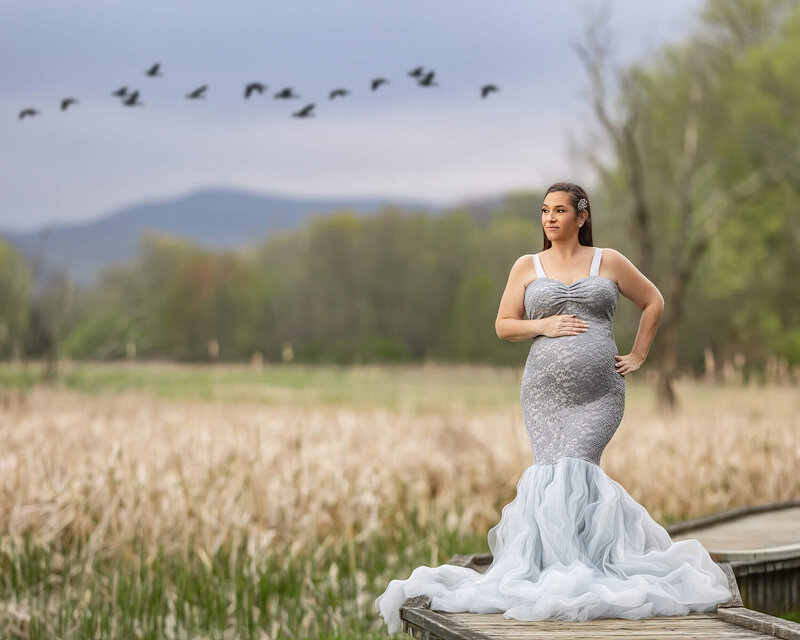 A pregnant woman in a grey lace and tulle gown is standing on a wooden walkway, holding her belly with the mountains and birds in the background