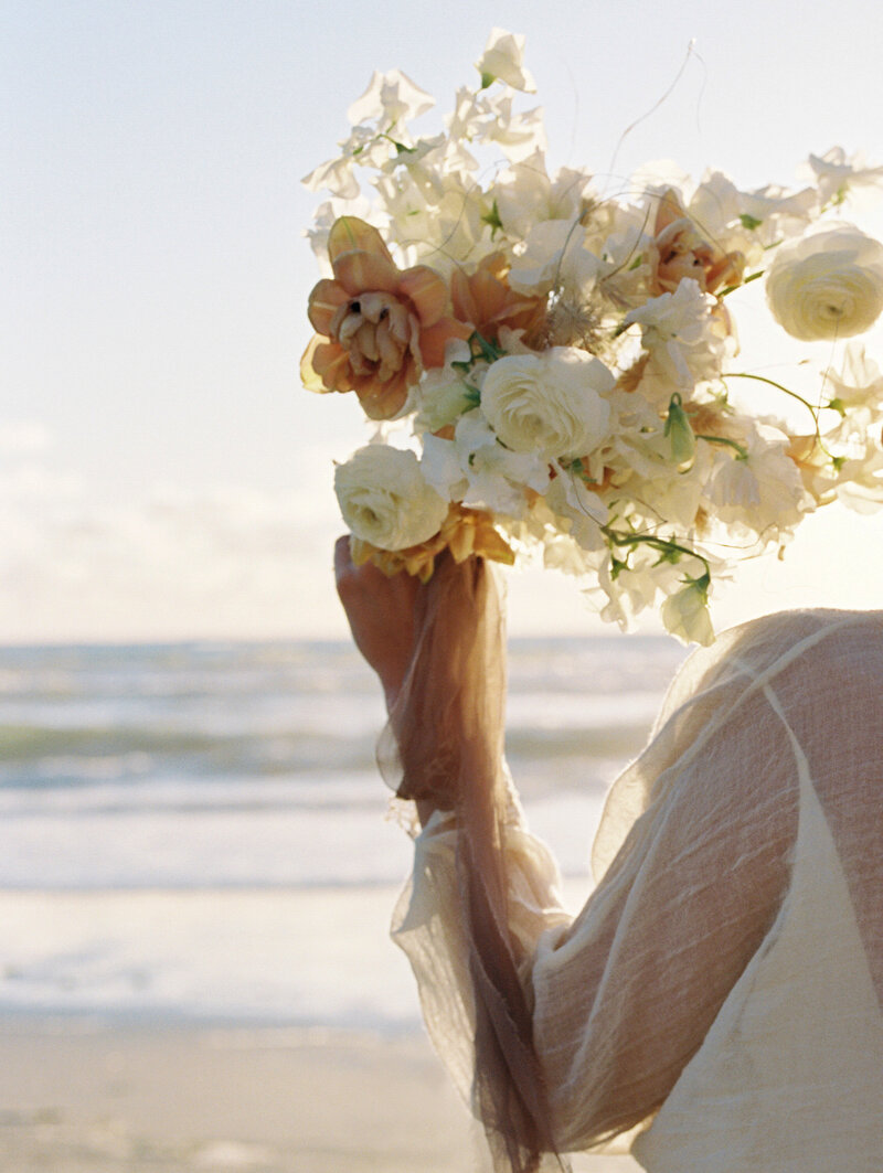 bride holds neutral wedding flowers with beach behind her
