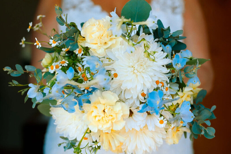 A close-up of a bridal bouquet featuring white chrysanthemums, soft yellow carnations, light blue flowers, daisies, and eucalyptus leaves, beautifully captured by a film photographer NJ, held by a person in a white lace dress.