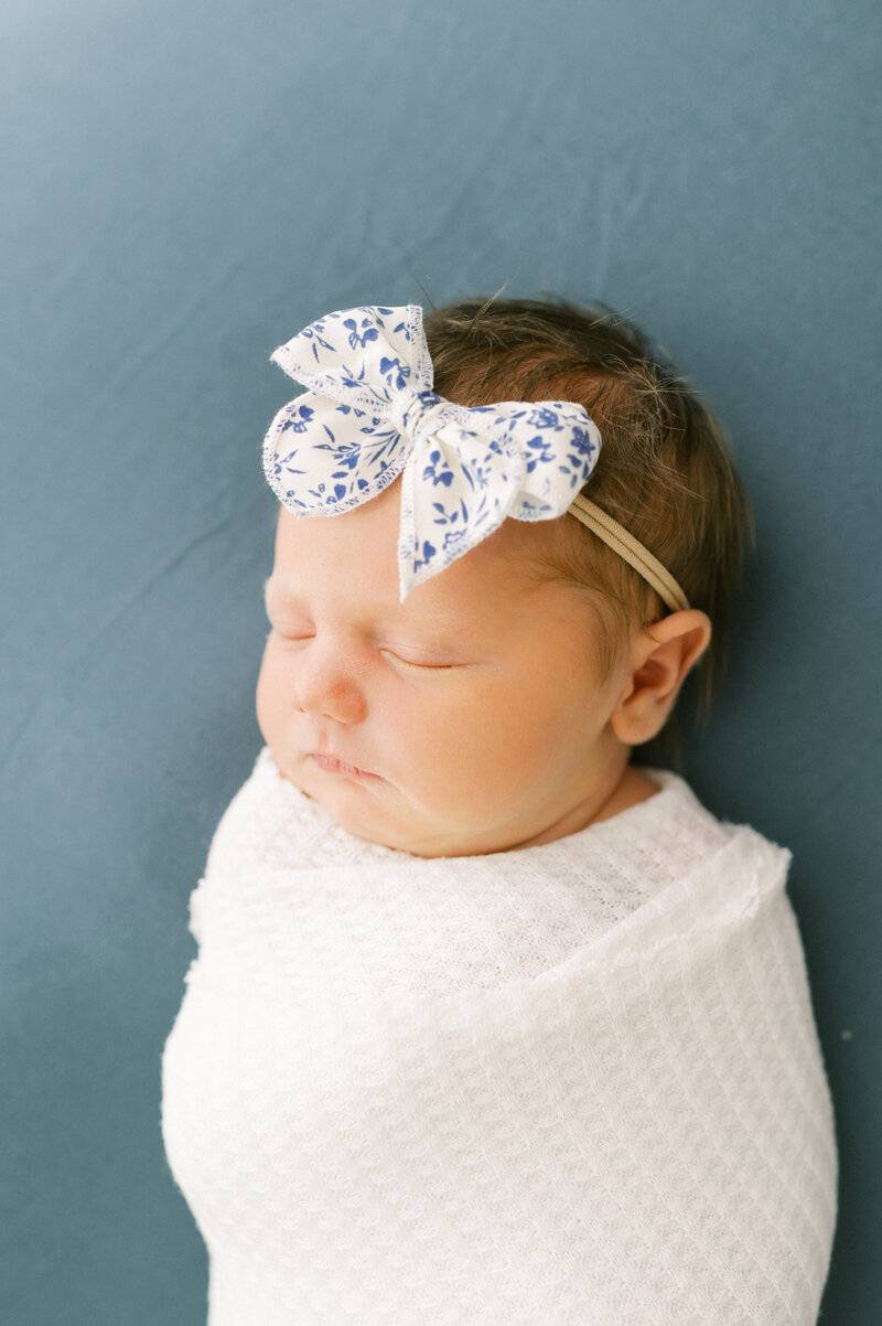Newborn girl with bow on her head swaddled up and sleeping on a teal background