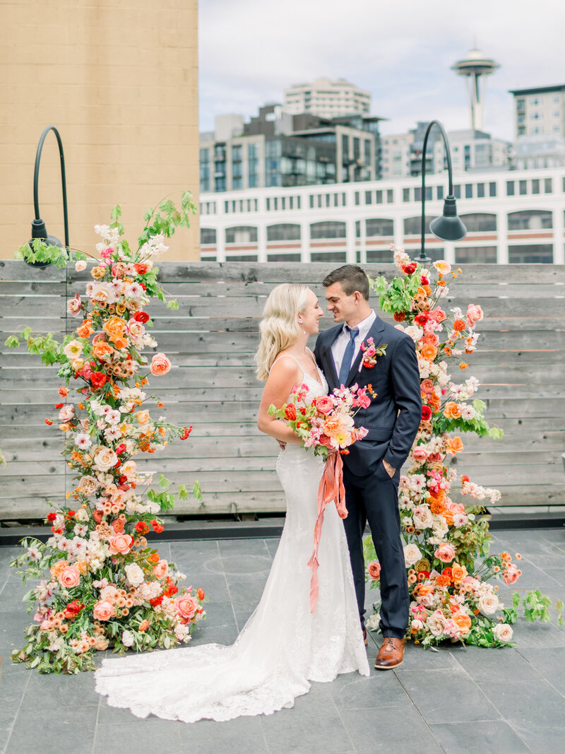 Photo of a bride and a groom standing a terrace overlooking the Seattle skyline and the space needle surrounded by bright boho flowers.