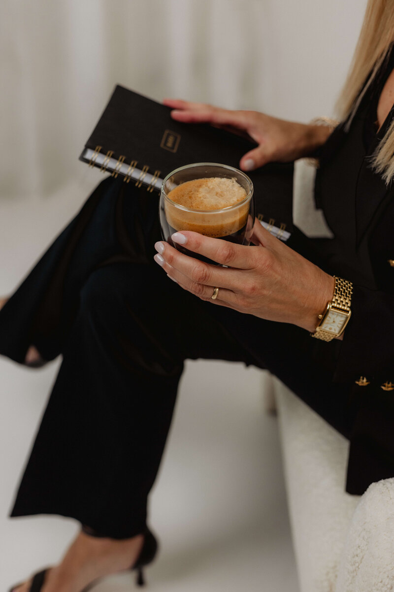 A women in a black suite holding onto a coffee and a book with her legs crossed.