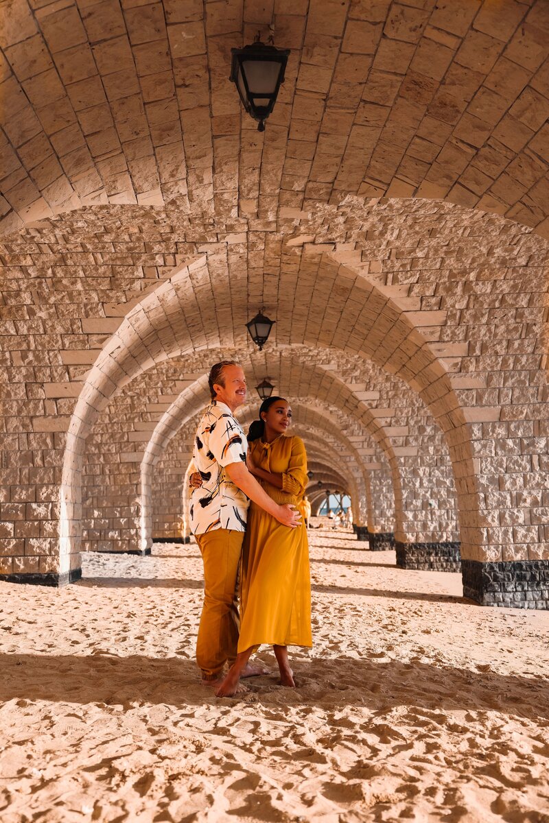 Voyagers Escape founders, Brett Schneider & Zee James, embrace underneath an arched stone walkway on a beach