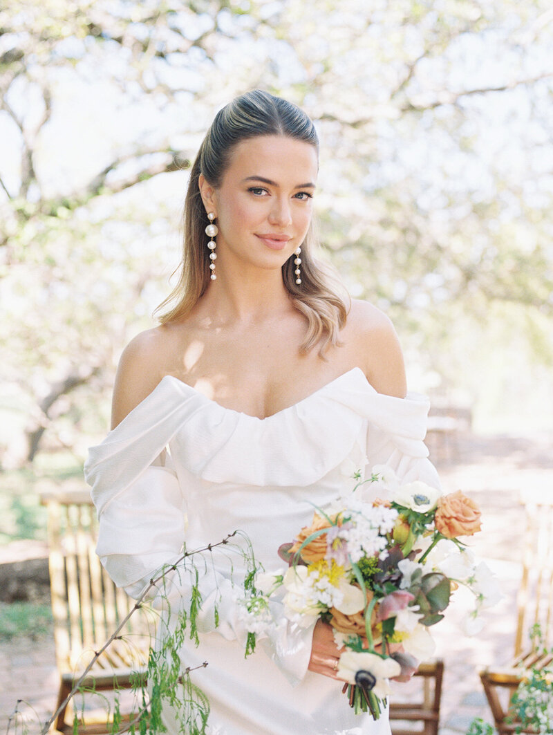 Destination wedding bride with pearl drop earrings in a white gown and pulled back hair smiling and holding a bouquet