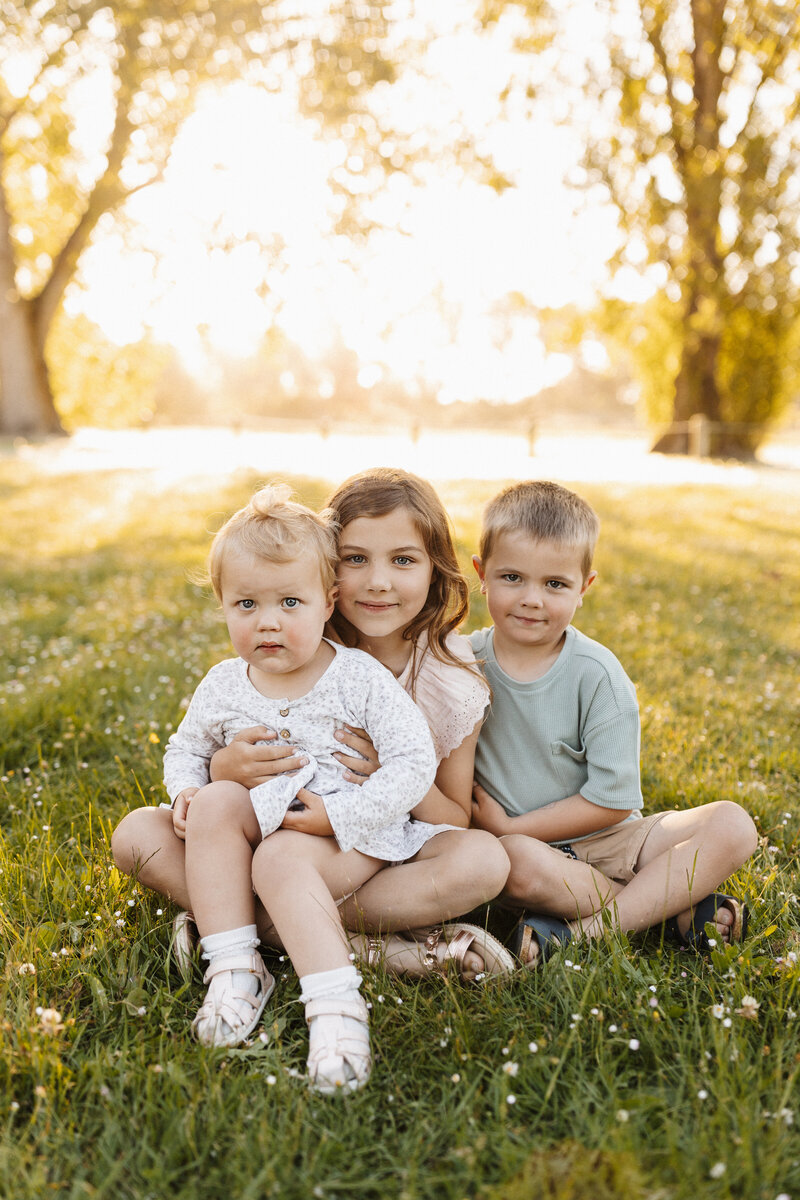 A set of three siblings sitting in the grass while the sunsets behind them during a christchurch family photoshoot with Christchurch photographer Lucy Jane Photography