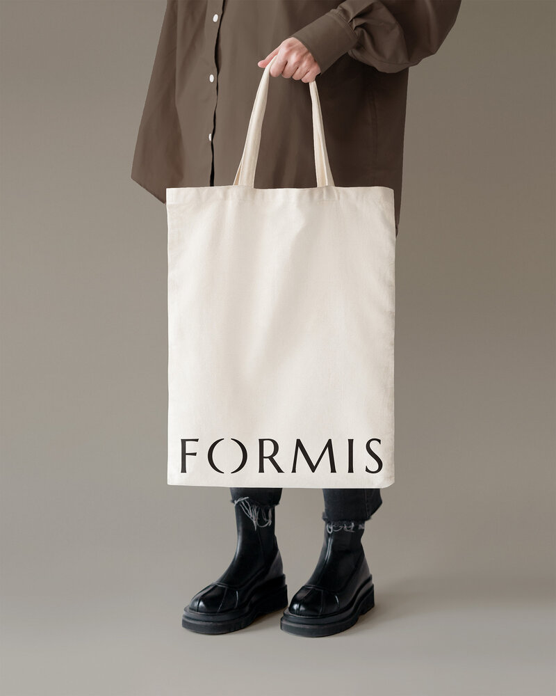 Crèmekleurige katoenen totebag met Formis logo, vastgehouden door persoon in donkere outfit