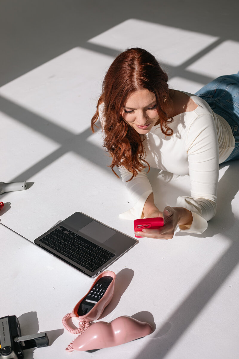 Mimi from Wed & Wilder lying on the floor in a white top and jeans, smiling at her red phone, with a laptop and a pink phone case beside her.