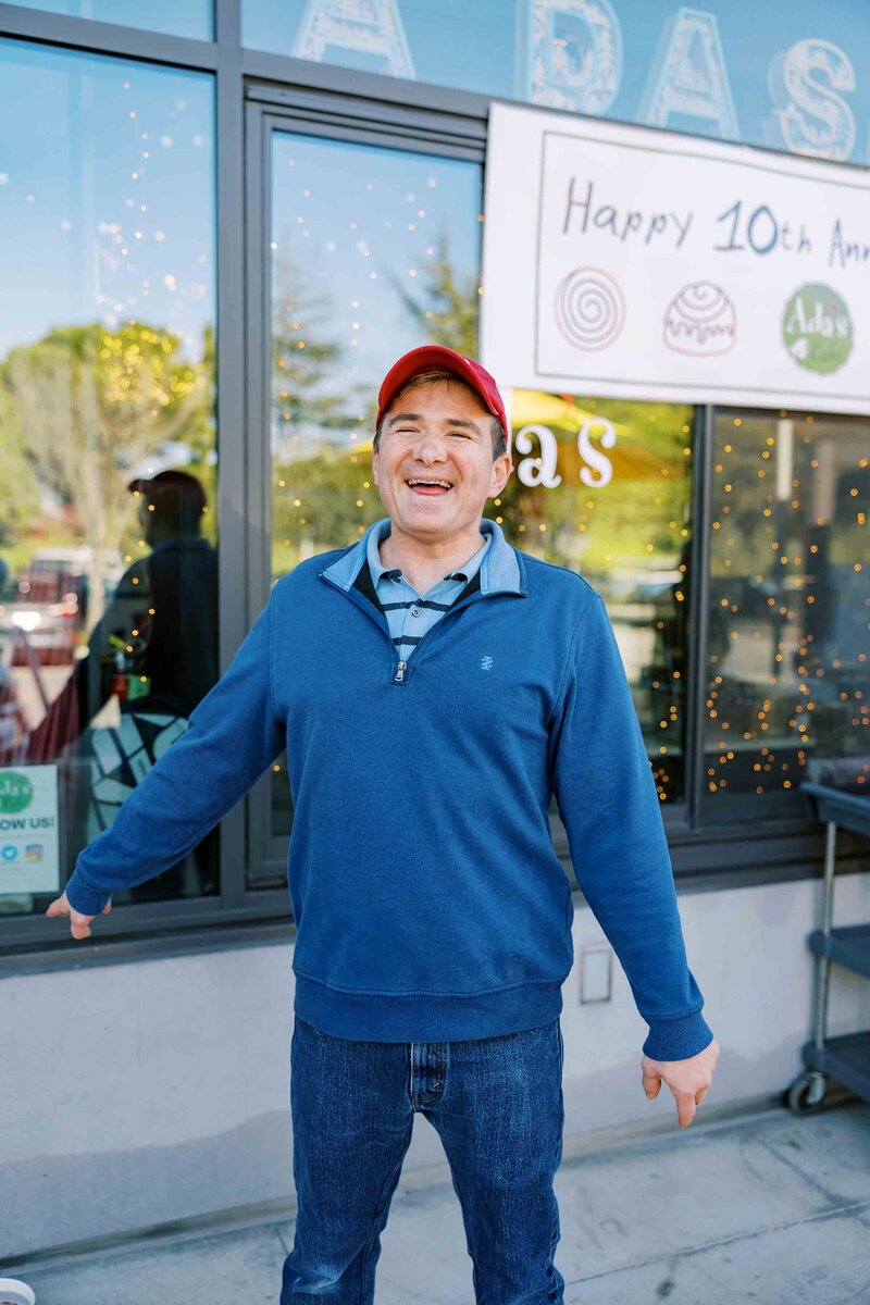 Man wearing a blue sweater, jeans, and a red baseball cap and smiling at the camera