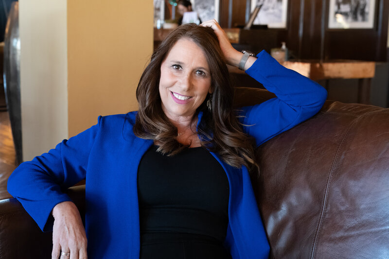 Smiling woman in a bright blue blazer lounging on a leather sofa in a warm indoor setting, photographed by Vyrl Photo, showcasing Tucson brand photography.