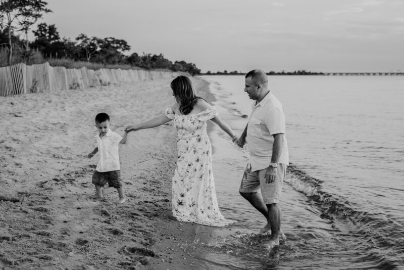 Black and white family photo of young son leading mom and dad from the ocean on the beach.
