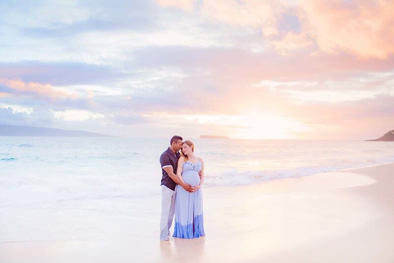Pregnant woman in blue maternity dress standing on the shoreline on Maui and being kissed by her husband during their Maternity portraits in Wailea with Love + Water