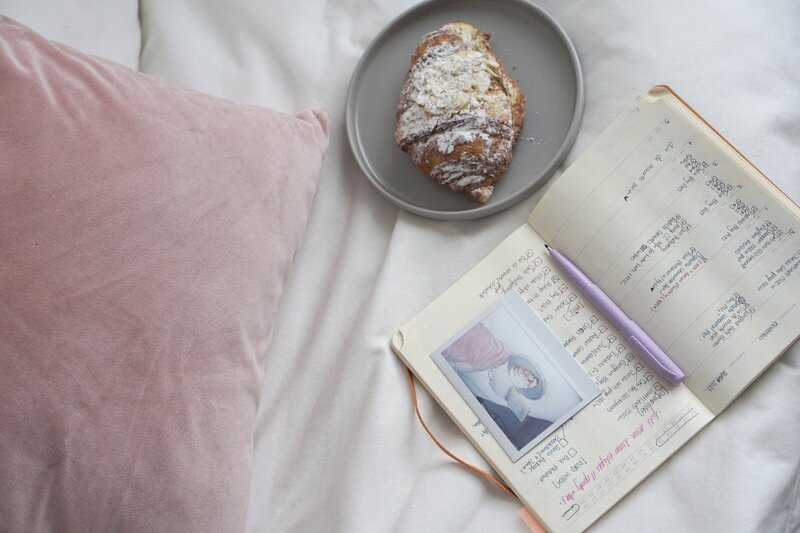 Overhead shot of a pink velvet pillow, an almond croissant, and a notebook open to a page with writing and a polaroud on top of the open pages.